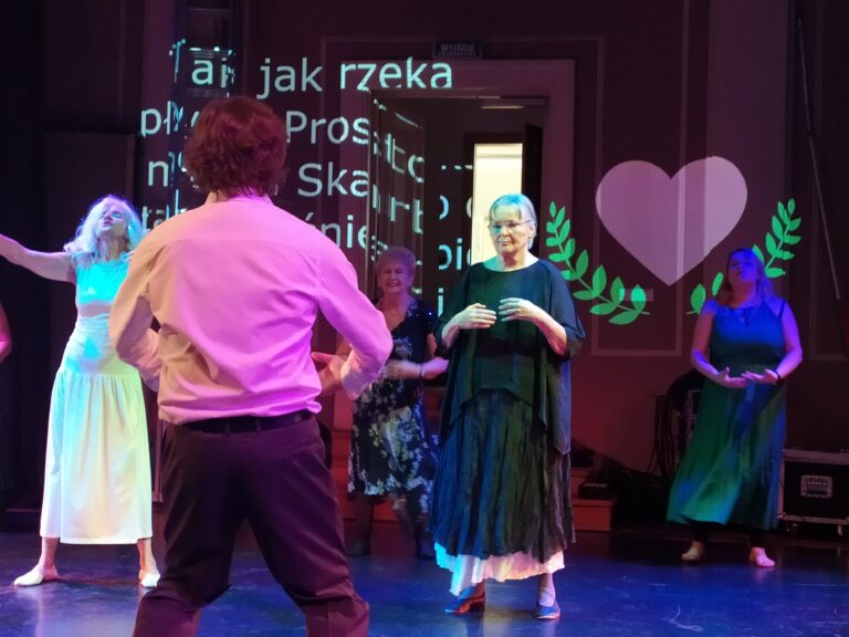 Kobiety przygotowujące się do tańca na scenie. W tle projekcja. Na pierwszym panie stojący tyłem mężczyzna prowadzący grupę.