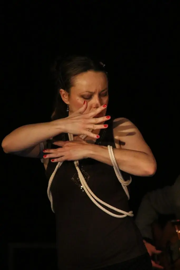 Tancerka flamenco w ekspresyjnej pozie z rękami przysłaniającymi dolną część twarzy i dekolt, ubrana w ciemnobrązowy top, z tułowiem oplecionym białą liną.